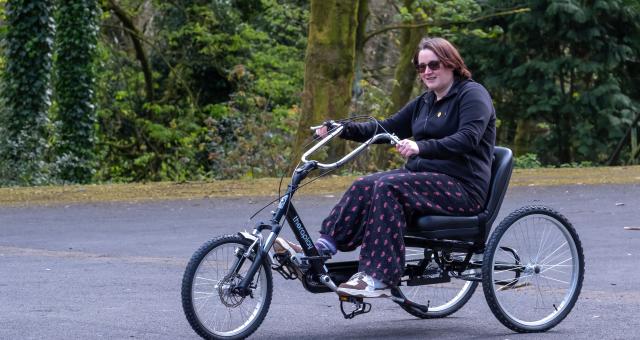 A woman is riding a tricycle in a park. She is wearing normal clothing and smiling