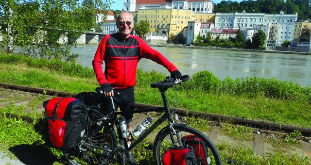 A man poses by a river with his bicycle. There are buildings in the background