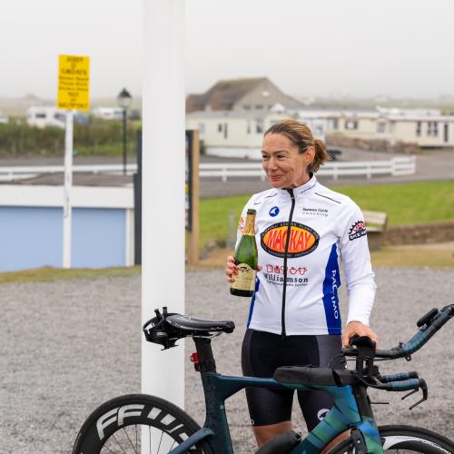 A woman is standing under a John o’ Groats sign. She has a Liv road bike with tri bars and is holding a bottle of champagne, celebrating after completing the Land’s End to John o’ Groats cycle ride.