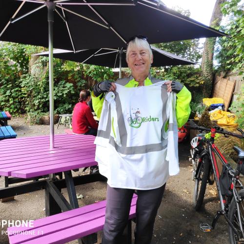 A woman is standing in a pub or restaurant garden. She is wearing cycling clothing and gloves. She is holding up a JoyRiders jacket