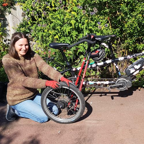 A woman is fixing a tyre on a cycle. She is wearing a brown sweater and black and red gloves. She has long brown hair. She is smiling at the camera