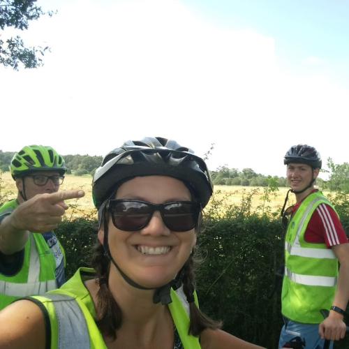 A selfie of three cyclists next to a hedge. The photo has been taken by a woman and behind her are two men, one to the left and one to the right. They are all wearing hi-vis jackets and cycling helmets