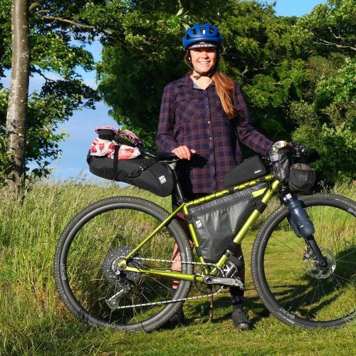 A woman is standing with a metallic green bike that is fully loaded with bags for bikepacking. She is wearing a blue cycling helmet and a check shirt. She is smiling at the camera
