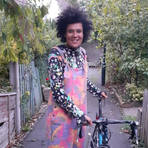 Belinda stands to the left of her bike on a path. She is wearing colourful dungarees and a black long-sleeved top.