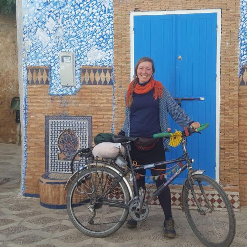 Eilidh smiles with her bike during one of her cycle adventures overseas. She is wearing a blue top and red scarf. Behind her is a colourful, historic building.  Her bike has a big yellow flower attached to the handlebars.