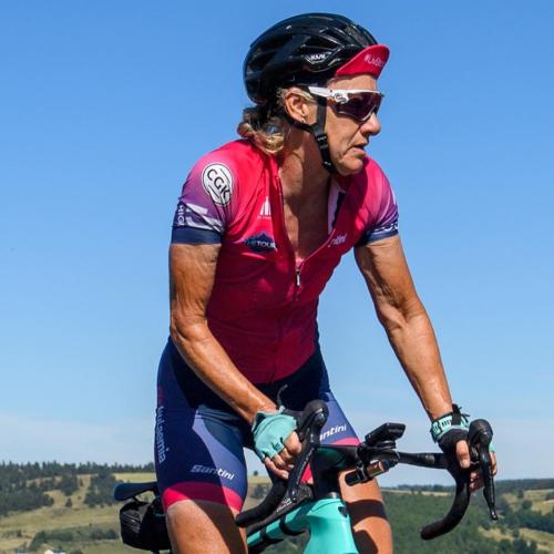 Jackie riding her blue bike in the countryside. She is wearing a pink jersey, blue shorts, black helmet and sunglasses.