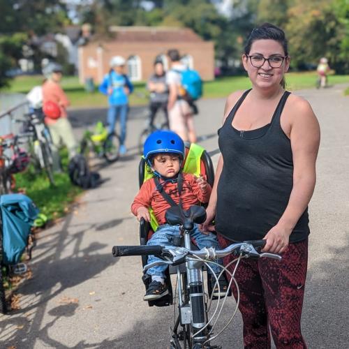 Kat stands to the right of her bike in a park. A small child is in a child’s seat at the back of the bike. Kat is wearing glasses, a black top and red patterned leggings.