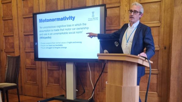 A man with grey hair and glasses, wearing a blue suit, is standing at a wooden lectern in a wood-panelled room. He is pointing at a large TV screen which has the word 'Motonormativity' on it