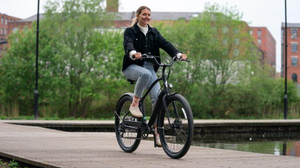 A woman smiles as she rides an electric bike along a wooden path beside a canal, with trees and red-brick buildings in the background.