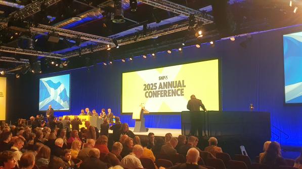 A large crowd of people sat around a stage with a panel of people sitting on it and a person standing to the right. There's a large screen that reads 'SNP 2025 annual conference'. There are also a couple of screens showing the St Andrew's Cross