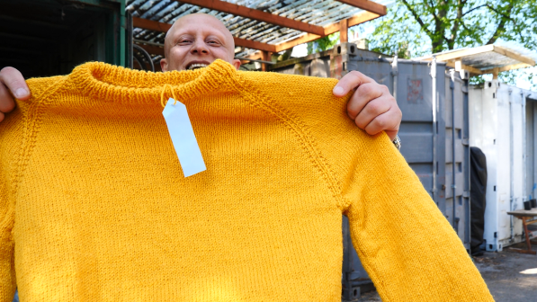 Mostafa smiles as he proudly holds up a bright yellow knitted jumper with a tag, standing outside near a workshop or storage area.