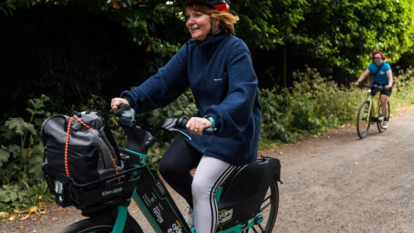 A woman rides an electric bike along a tree-lined gravel path, smiling and wearing a helmet. Two other cyclists follow behind her.