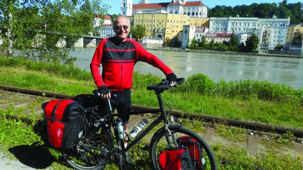A man poses by a river with his bicycle. There are buildings in the background