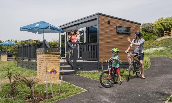 A family waving at each other, next to a glamping pod and on bikes. 