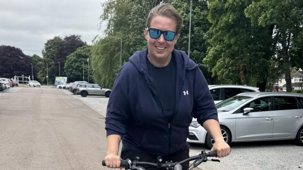 A woman in sunglasses and a navy hoodie smiles as she rides an e-bike through a car park. She appears confident and relaxed, enjoying the ride on a cloudy day in Luton.