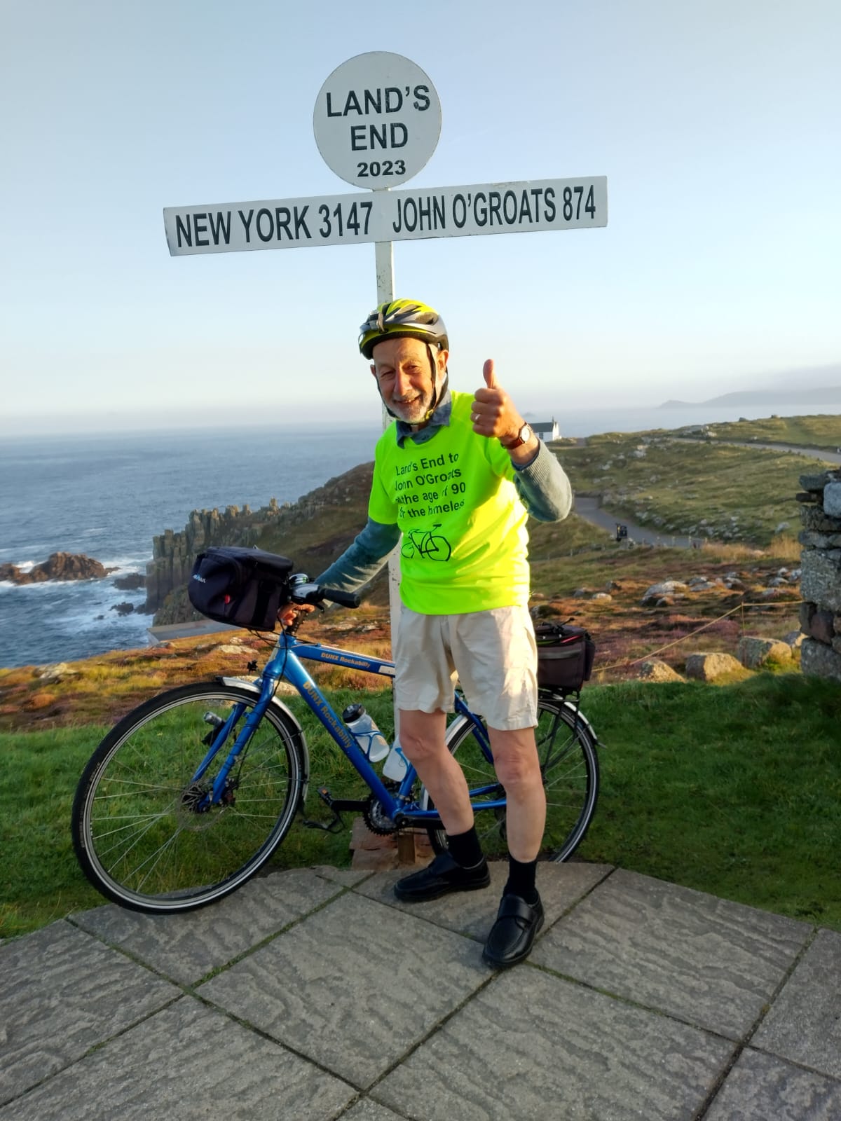 A man stands with a bike under the Land’s End signpost. He is wearing a bright yellow T-shirt and giving a thumbs up to the camera