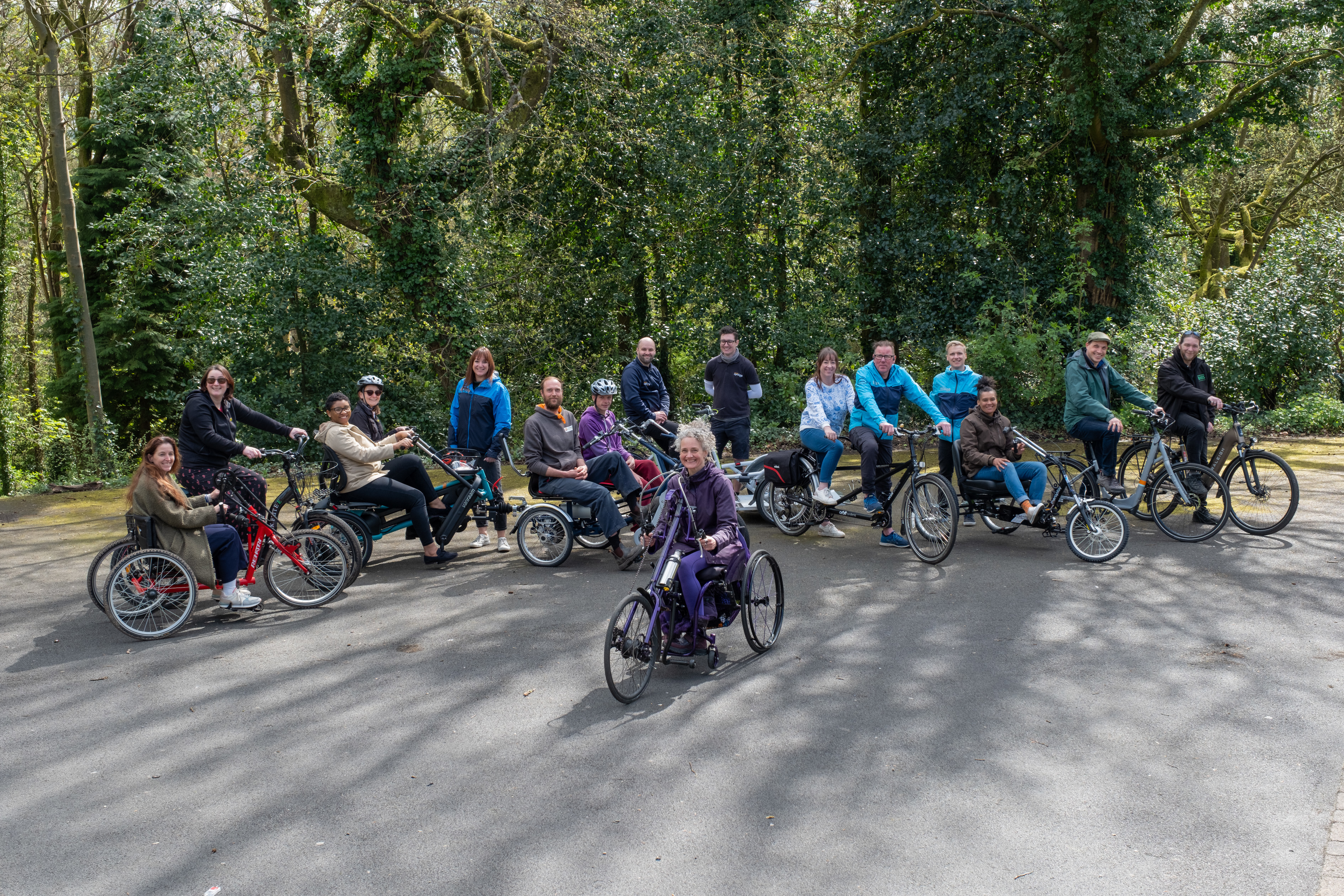 A group of adults in a park on a variety of non-standard cycles, tandems, e-assist cycles, handcycles and tricycles