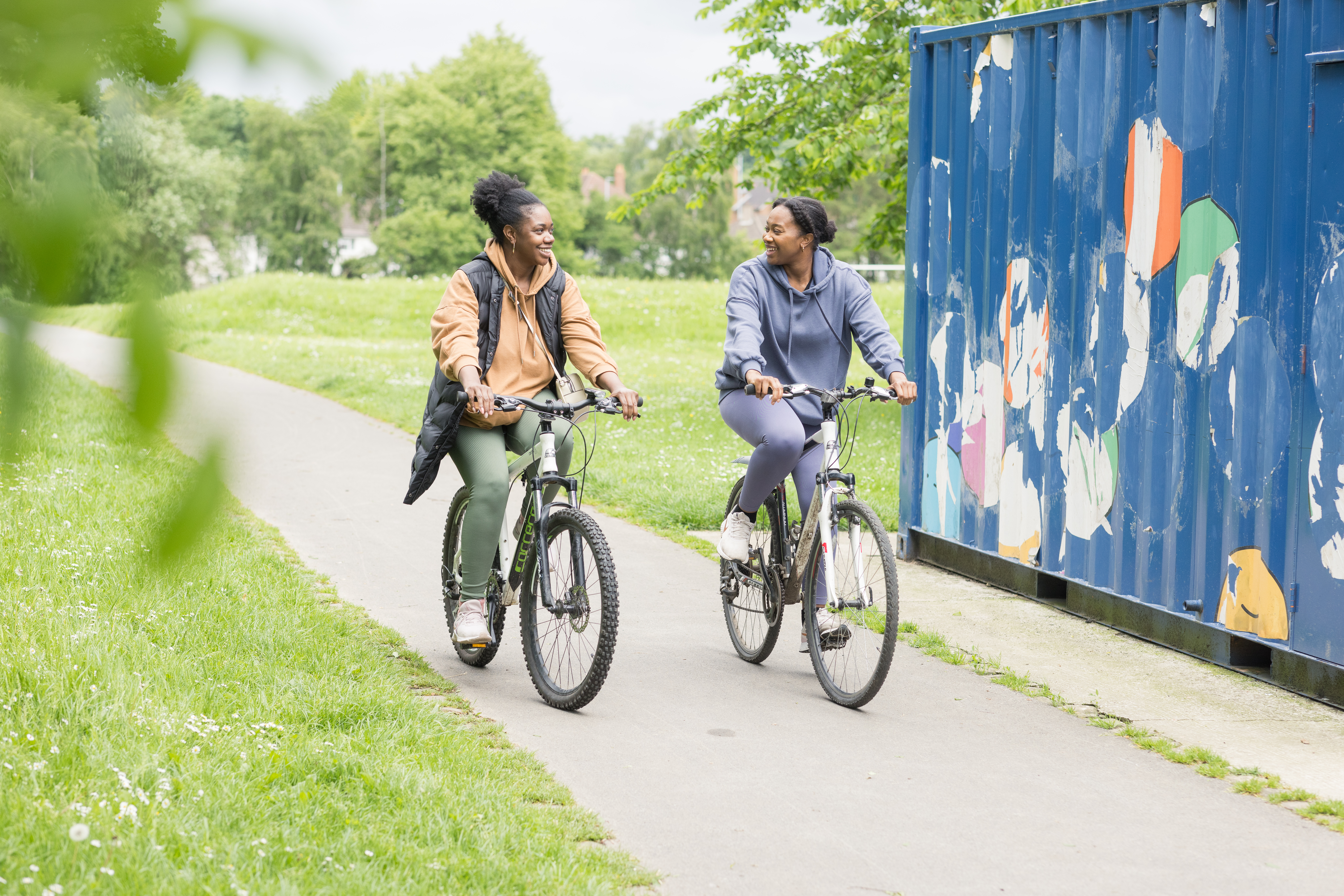 Two women cycling on a path in a park with a shipping container on their left