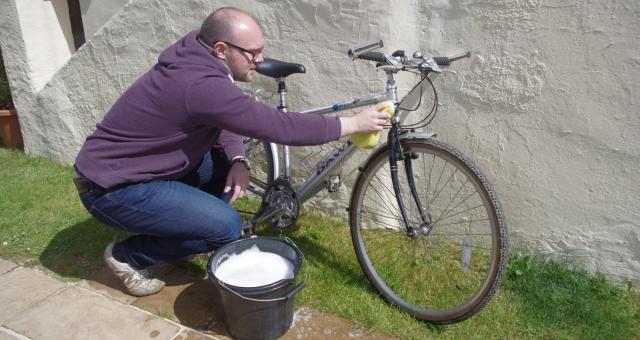 A man wearing jeans and a purple hoodie is kneeling down to clean a silver bike that's leaning against a white wall. He's using a big sponge and there's a bucket of soapy water next to him