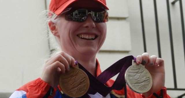 Paralympic cyclist Aileen McGlynn holding her silver and bronze medals won at the 2012 Summer Paralympic Games. She is wearing Team GB colours, a red peaked cap and cycling sunglasses.  Photo: The Rambling Man and Kim Ratcliffe of Think Equestrian under the Creative Commons Attribution-Share Alike 3.0 Unported license. https://creativecommons.org/licenses/by-sa/3.0/deed.en