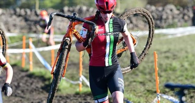 Aisling Charlesworth carries her cyclocross bike across a muddy section of a race. She is wearing red, white, green and black Fibrax Wrexham Roads Club kit, a helmet and sunglasses. She is covered in mud!