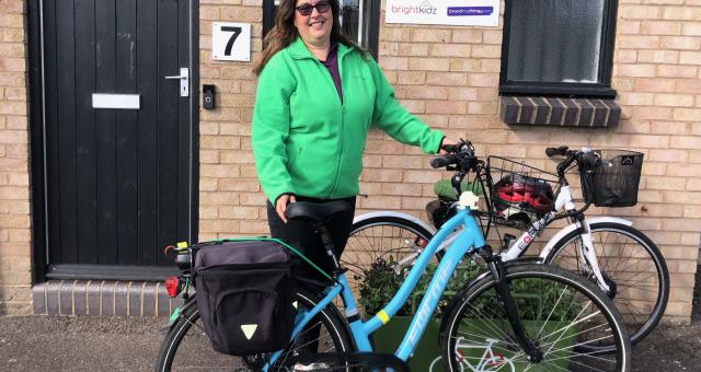 Alison Holland is standing outside a brick building. She is holding a blue hybrid bike with a front basket and rear panniers. She is wearing a green fleece and black trousers. She has long brown hair and sunglasses. Behind her is another bike, a white hybrid.