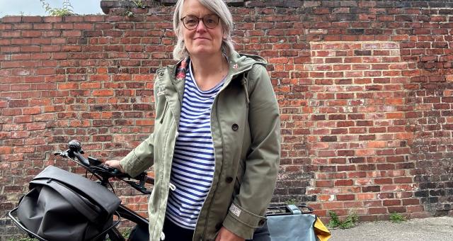 Alison Stenning is standing in front of a brick wall, holding her black hybrid bike behind her. It has a front basket, rear pannier and frame bag. She is wearing a green jacket, stripey top and black jeans.
