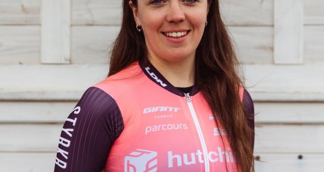 Amy Marks is standing in front of a white-painted wooden fence. She is wearing a purple and pink short-sleeved cycling jersey. She has long brown hair and cycling sunglasses on her head.
