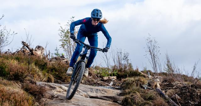 Anna Riddell is riding a mountain bike down a technical off-road trail. She is wearing blue mountain biking kit and helmet and cycling sunglasses. She is smiling.