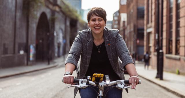 Beccy Marston is riding along an urban street. You can just see the flat handlebar of her bike. She is wearing a grey jacket with white check pattern, black top and blue jeans. There is a rainbow badge on the lapel of the jacket. Her smartphone is attached to her handlebar. She is smiling at the camera.