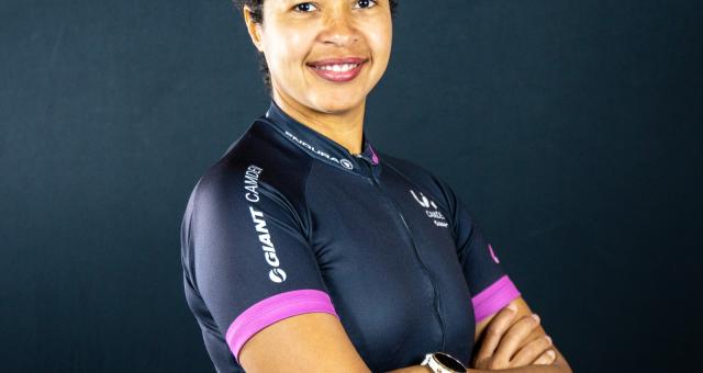 A portrait of Bianca Fernandez. She is standing with arms folded against a blue-grey background. She is wearing a dark blue cycling jersey with a multicoloured pattern around the bottom and purple cuffs.