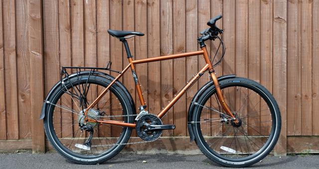 A copper-coloured flat-bar touring bike with black rear rack and mudguards leaning against a wooden fence