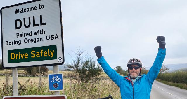 Mark Wedgewood standing next to the sign for Dull Scotland with his arms in the air and his bike leaning beside him