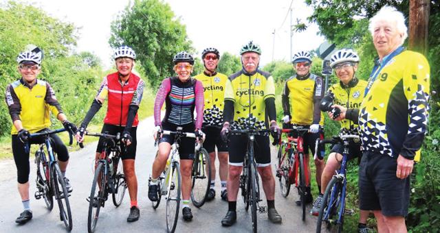 A mixed group of people are all standing with their bikes. They are mostly wearing yellow and black club kit. A couple of people are in non-club cycling kit. They are wearing helmets. They are all smiling at the camera