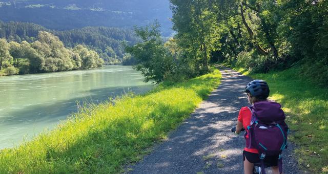 A child is cycling along a gravel track alongside a river. There are mountains in the background and the track is lined with trees. The child is on a mountain bike and wearing shorts and T-shirt, with a cycle helmet and backpack