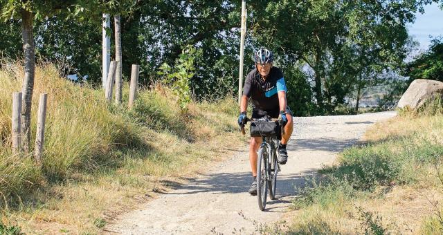 A man is riding along a gravel track through countryside. He's riding a touring bike with a handlebar bag on the front. He's wearing blue and black cycling short-sleeved jersey and shorts, a cycle helmet and glasses