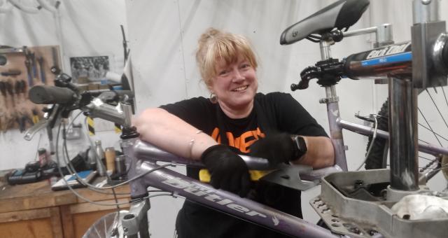 Cath Palgrave is working on a light metallic mauve Claud Butler mountain bike in a mechanics’ workshop. The bike is clamped to a work stand. Cath is wearing a black T-shirt and black gloves. She has blonde hair and hoop earrings. She is smiling at the camera.