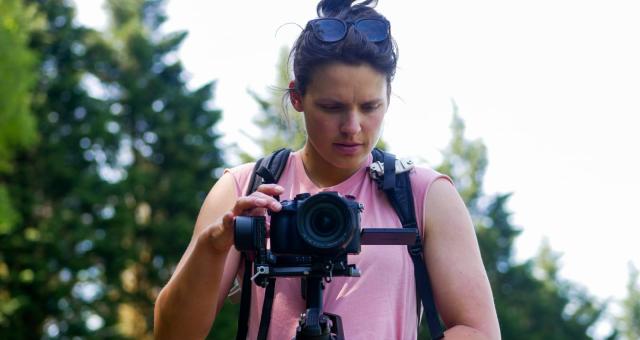 Filmmaker Catherine Dunn peering into her camera in a woodland. She is wearing a pink sleeveless t-shirt and a rucksack.