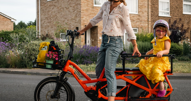 Caz Conneller is sitting astride an orange longtail cargo bike. She is wearing three-quarter-length blue jeans and a shirt. She has a helmet on. A little girl is sitting on the back of the bike. She is wearing a yellow party dress and patterned helmet. Two bags and a pot plant are in the basket at the front.