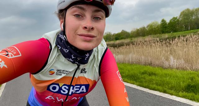 Charlotte Broughton has taken a selfie as she rides along a road. She is wearing race kit: a UCI Women’s Continental Team jersey and red arm warmers, a blue buff and white helmet. She has cycling sunglasses tucked into her helmet.
