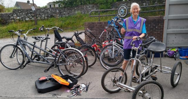 Christine Fisher is standing surrounded by lots of bikes, including tandems, trikes and quads. There is a big toolbox open on the ground and she is holding up a tool with her other hand on a silver quad bike.
