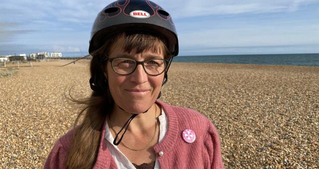 Cicely Lloyd is standing on a pebble beach with the sea in the background. She is wearing a black Bell helmet, a pink cardigan and white shirt. She has a BMW Cycle Club badge on her cardigan. She has long brown hair and glasses. She is smiling at the camera.