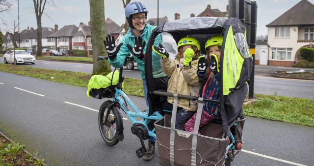 A woman is standing next to a pale blue cargo bike on a paved cycle path that is separated from the road. There are two young children in the cargo box. They are all holding up their hands to the camera to show their gloves which have smiley faces on them