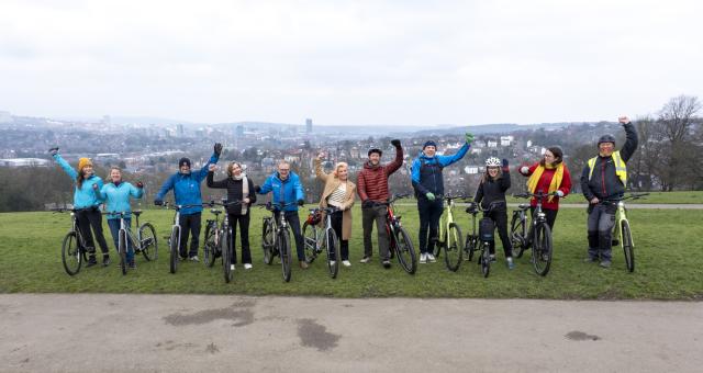 A row of people stand with bikes on a hill with a view over Sheffield in the background. They are smiling and waving at the camera. In the centre is Louise Haigh, Secretary of State for Transport