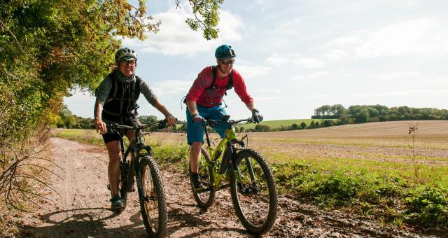 Two people are riding mountain bikes on a muddy trail in the countryside. They are wearing shorts and T-shirts and cycle helmets