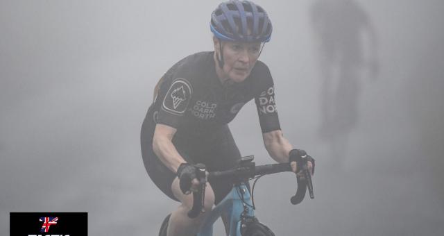 Deborah John riding a road bike in full cycling gear. She has a focused look on her face and is surrounded by a hazy mist, which makes the rider behind her hard to see, but gives the photo and eerie but beautiful quality.