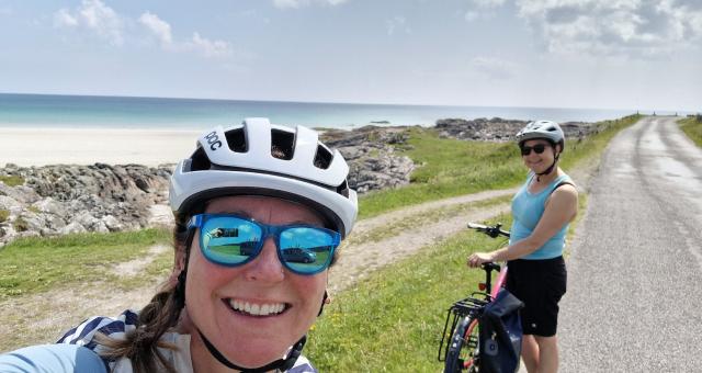 Diane Clayton has taken a selfie. She is wearing a white cycle helmet and cycling sunglasses. Another cyclist is behind her, and in the background is a dry stone wall and beyond that the sea.