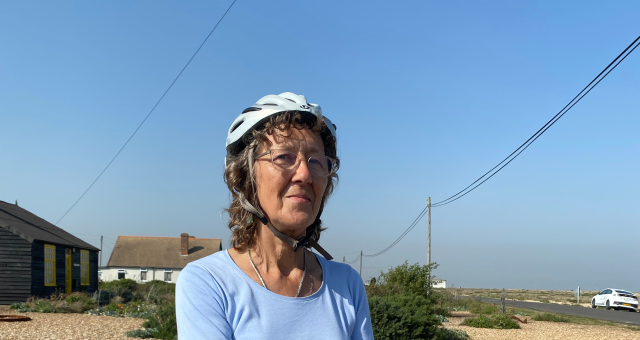 Eilidh Murray posing with her bike, wearing a helmet, light blue T-shirt and darker, smoky blue shorts. She is at Dungeness, in front of Prospect Cottage, the former house of artist Derek Jarman