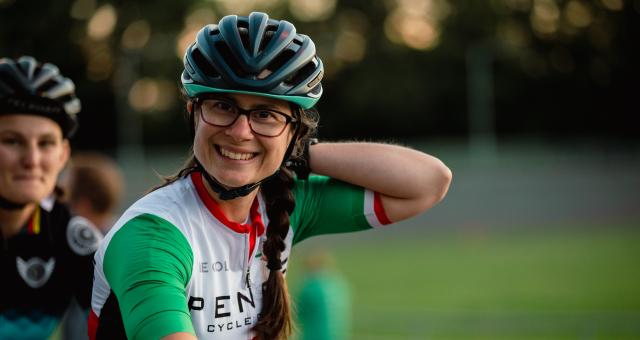 Elisabetta Motta is wearing brown rimmed glasses a dark green helmet and Lycra cycling top and is smiling with her left arm raised in the air reaching for her back