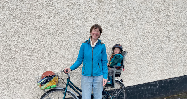 Emily Ryder is standing by a white wall with her cycle leaning up against the wall. She has an assortment of things in her front basket and her young child is sitting in the rear child seat